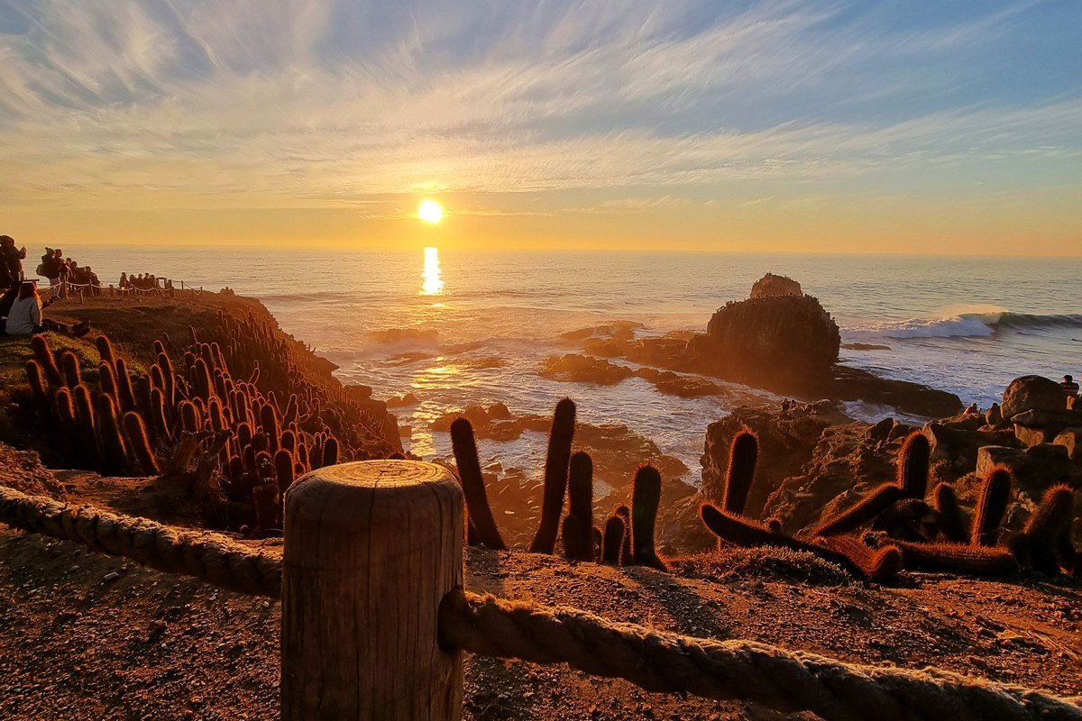 Moonstone Beach in Cambria at sunset, smooth agates and driftwood in the foreground, the bluff boardwalk visible to the north