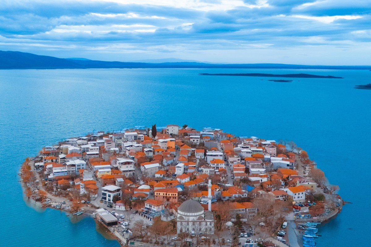 Aerial view at golden hour over a whitewashed clifftop village, turquoise water catching the last direct light below, a single fishing boat anchored in a cove to the lower right. Shoot from elevation to compress the village geometry against the sea. Avoid midday.