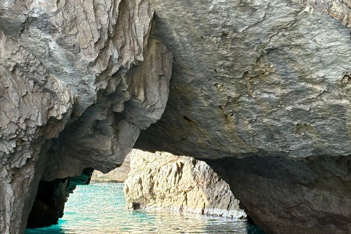 Low-angle shot from a kayak inside a Marettimo sea cave, sunlight refracting through shallow turquoise water onto the cave ceiling above. A limestone arch frames the open sea behind. Shot in the first two hours after sunrise before direct light bleaches the color from the water.