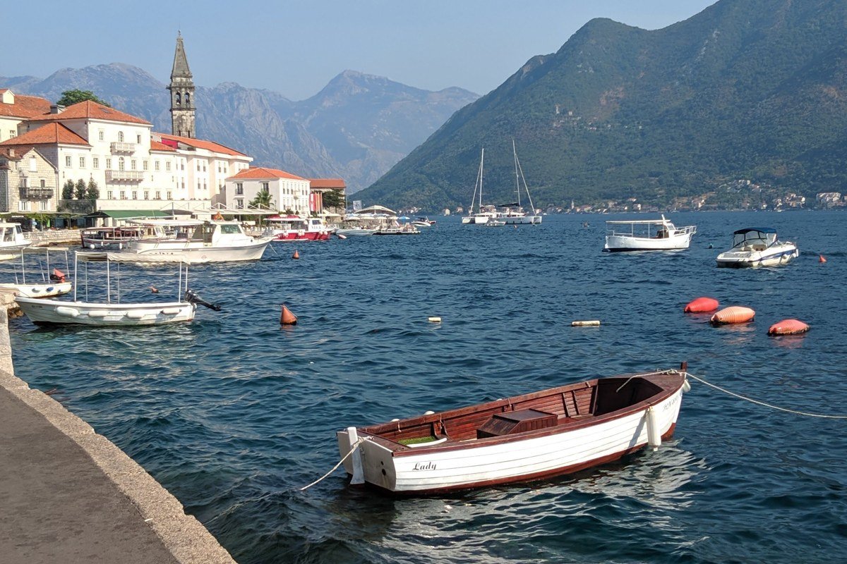 Kotor Bay at dawn, shot from a boat or kayak on the water facing the old town. The Venetian walls rise directly from the waterline. The Dinaric Alps reflect in the still early-morning bay. The fortification line above the town is visible on the hillside. Shoot before 7 a.m. in summer for flat water and warm raking light on the limestone.