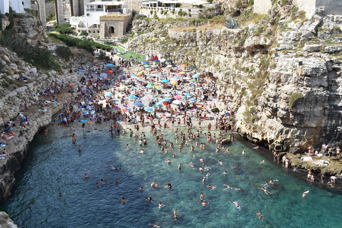 Lama Monachile cove, Polignano a Mare, shot from the Roman bridge looking directly down into the cove. The two limestone cliff walls frame the frame symmetrically. The water is clear enough to show the rocky seabed in the shallows. Shoot at midday in summer when the sun is directly overhead and the water color is at its most saturated. Include the bridge railing in the foreground for depth.