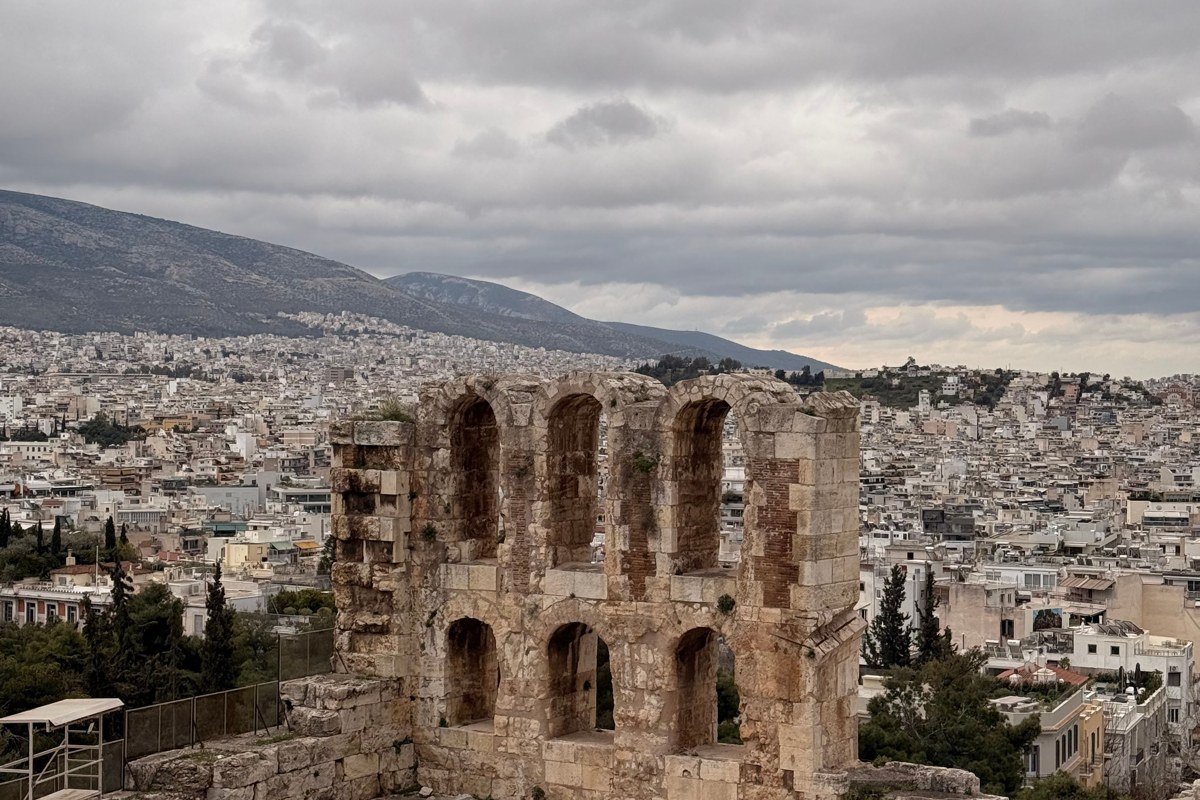 The ancient Greek-Roman theater at Taormina, shot from the upper seats looking through the surviving stone archways toward the stage. Mount Etna is partially in cloud on the left horizon, the Ionian Sea filling the right two-thirds of the background. Shoot in the last ninety minutes of afternoon light when the stone warms to amber and the sea turns silver-blue. Avoid shooting in the heat of the day when the contrast is too harsh for detail in both stone and water.