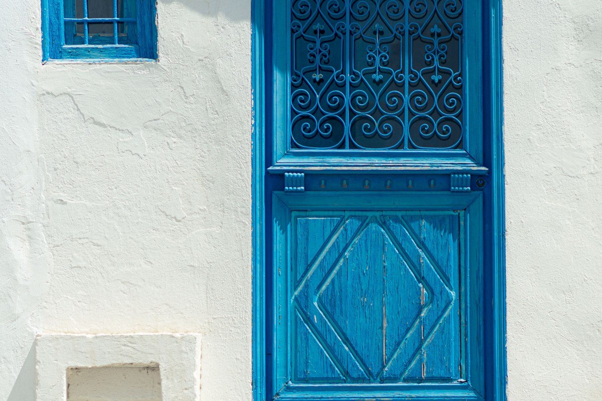 A Sidi Bou Said doorway in full shade, the cobalt-blue ironwork grille and frame set against a bright white wall. A terracotta pot of geraniums at the base left. The Gulf of Tunis is visible through a gap in the buildings in the far background. Shoot at 50mm to avoid distortion on the architectural geometry. The most graphic shots are in the two hours around midday when the shade is densest and the white walls hold detail without blowing out.