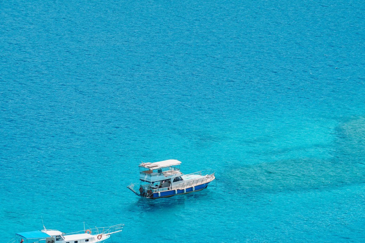 Kas harbor at early morning, shot from the hillside above the main square looking down over the rooftops toward the water. Kastellorizo island is visible on the horizon across the strait, its own small harbor distinguishable on a clear day. Bougainvillea in the foreground right. The Lycian rock tomb is partially visible at the left cliff edge. Shoot before 8 a.m. for flat water and before the day-trip boat traffic begins.