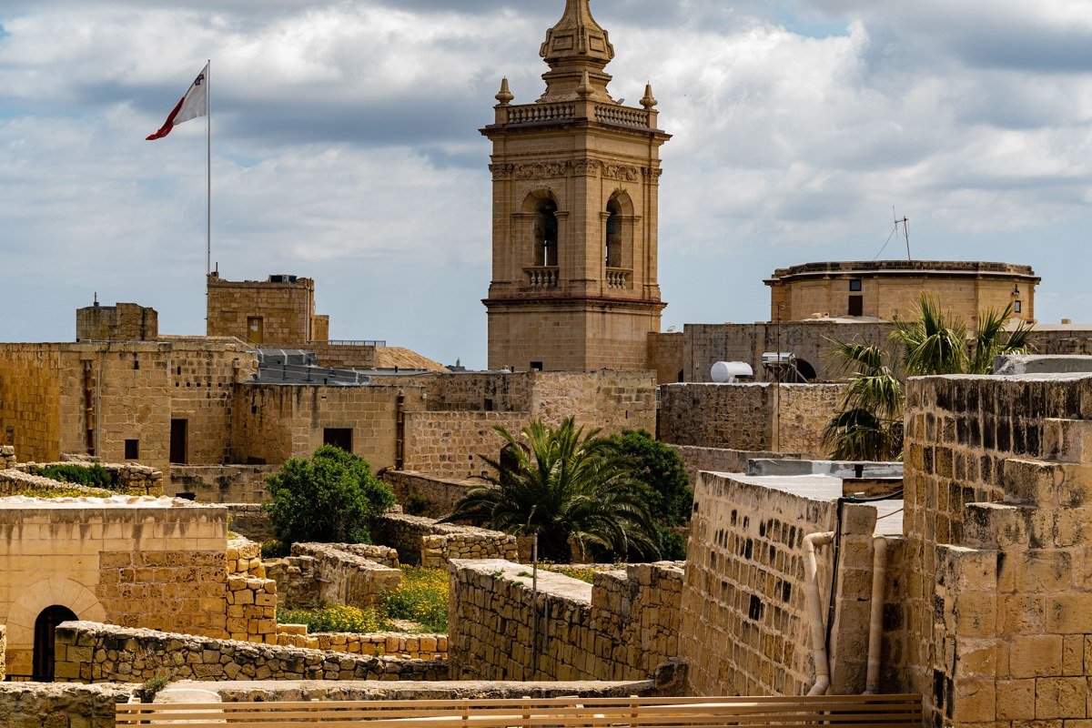 The Grand Harbour from the Upper Barrakka Gardens bastion wall, shot at golden hour looking southeast. The limestone fortifications in the foreground shift from cream to deep amber in the last thirty minutes of direct light. The Three Cities, Vittoriosa, Senglea, and Cospicua, are visible across the harbor with their own fortification lines intact. Shoot at f/11 to hold detail from the near fortifications to the far shoreline. The harbor reflections are sharpest in the fifteen minutes before direct sunlight leaves the water surface.