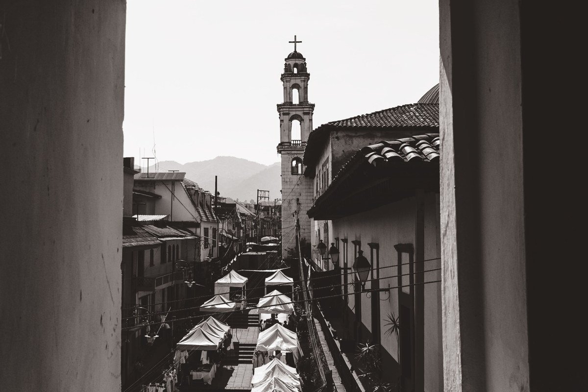 Llica colonial church facade in flat midday light, mud-brick bell tower casting hard shadow across dusty plaza, two women in pollera skirts visible at left edge of frame
