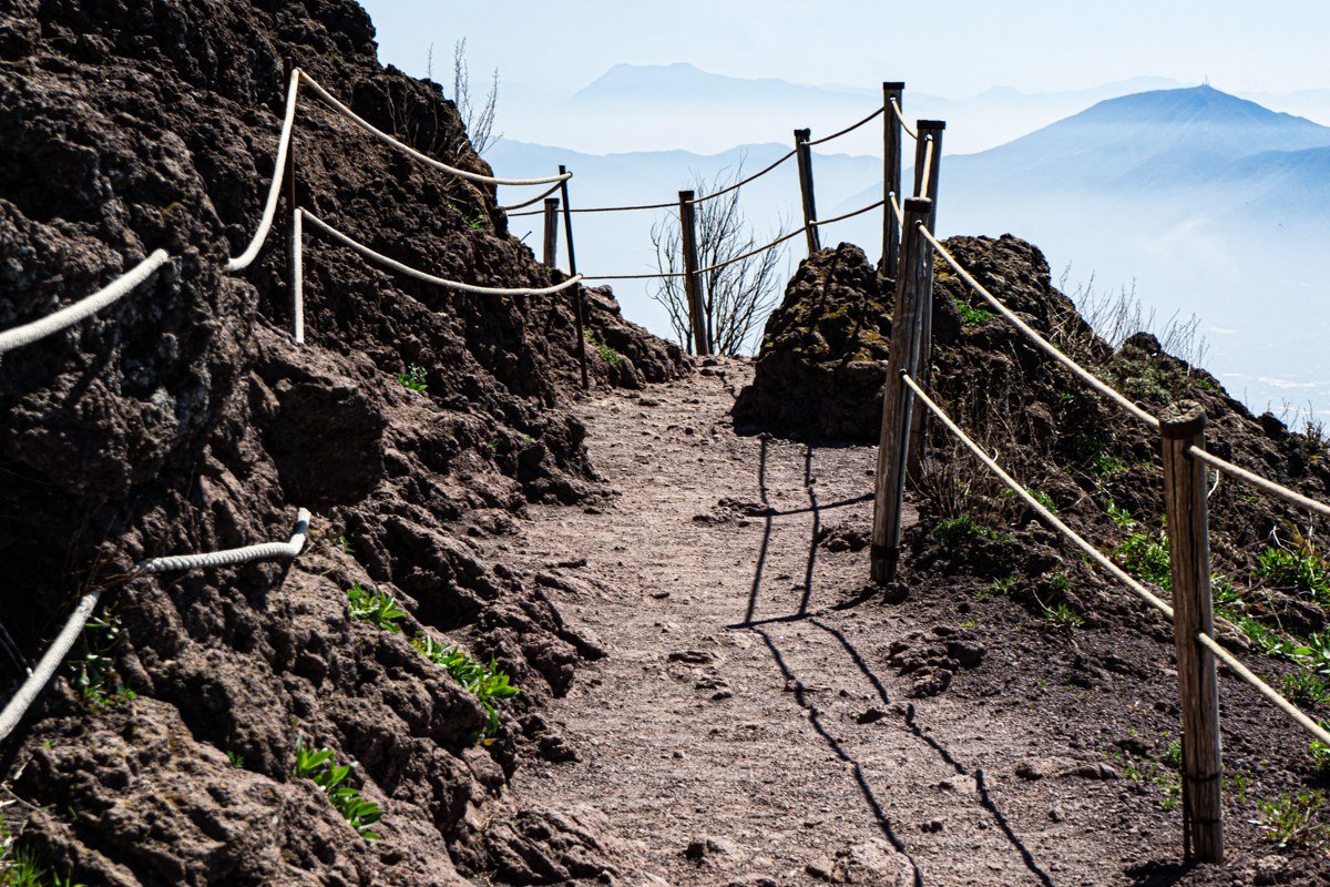 Steep rocky path above Coquesa village, Tunupa volcano summit in soft afternoon haze above, amber light catching the path's loose stones and a wooden directional marker
