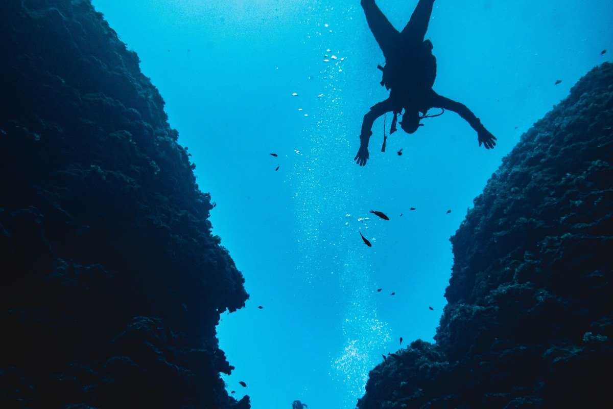 Shoot vertical from a low boat angle at dawn, Kadidiri Island silhouette, with a diver entering the water in the foreground. Drone for the archipelago overview shot only; keep dive imagery at water level.