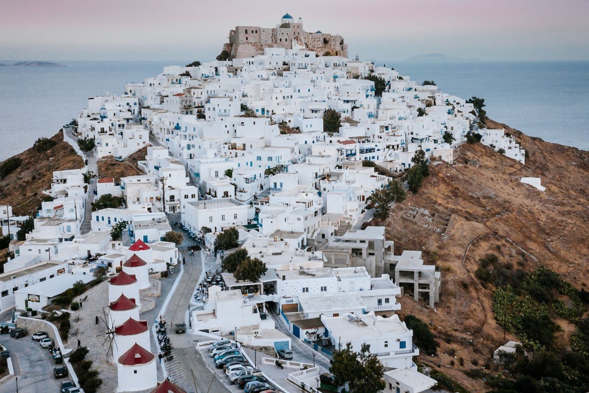 aerial shot at golden hour, whitewashed cube village on clifftop, cobalt Aegean in background, wild scrub hillside in foreground, no visible tourist infrastructure