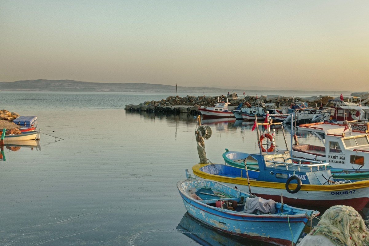 Livadia bay at mid-morning, turquoise water in foreground, three pastel fishing boats moored close to shore, small white chapel on left edge of frame, bougainvillea on waterfront visible behind boats