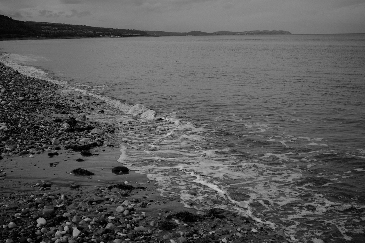 Nas beach at low sun, river mouth flowing over dark pebbles into sea in foreground, rocky headland with ancient temple ruins visible upper right, amber light on water surface
