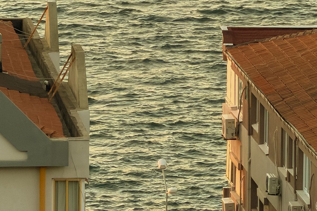shot from the mule path above Nimborio at golden hour, terracotta and ochre neoclassical facades in foreground, harbor water reflecting warm light below, Turkish coast faintly visible as a dark line across the strait in background