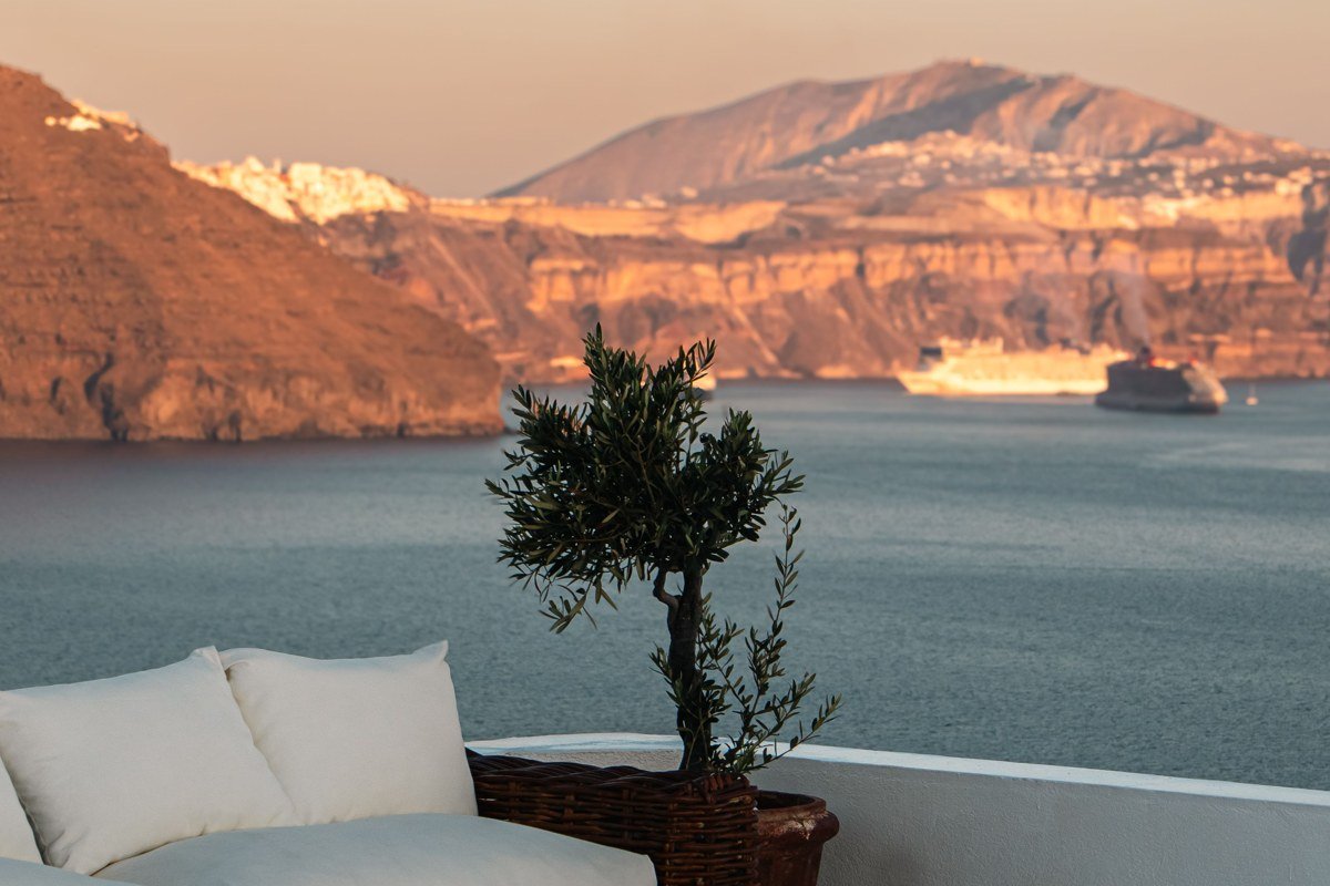 Manolas village taverna terrace at sunset, caldera visible below and beyond the terrace wall, Santorini's Oia visible as a white ribbon across the water in the far distance, shot over a carafe of wine in foreground, warm amber light