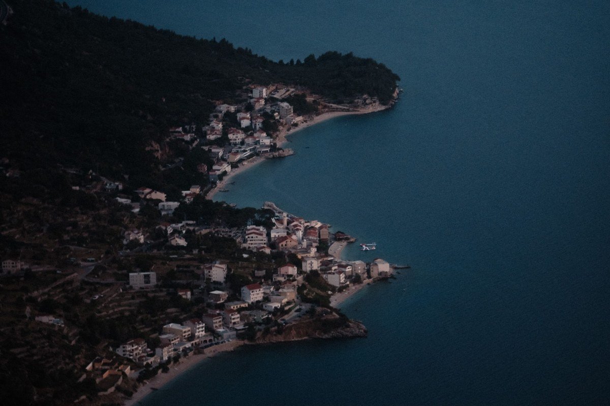 Meganisi Spartochori village at dusk, whitewashed and pastel-painted houses stepping down a hillside toward a dark Ionian sea, a single lit taverna visible at street level, pine trees framing the right edge of frame