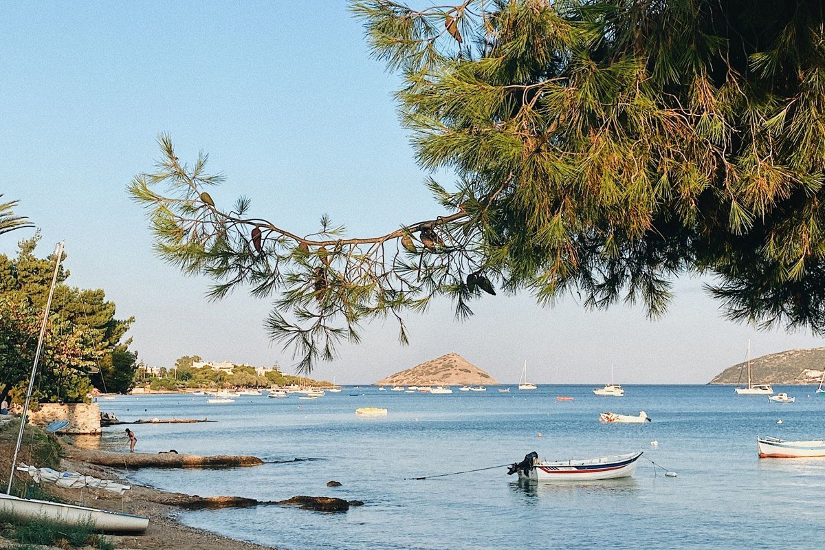 Agistri Skala harbour at late morning, pine trees coming down to waterfront on left, small blue and white caique moored at the quay, turquoise Saronic water in foreground, Athens mainland visible as a low grey line across the water behind