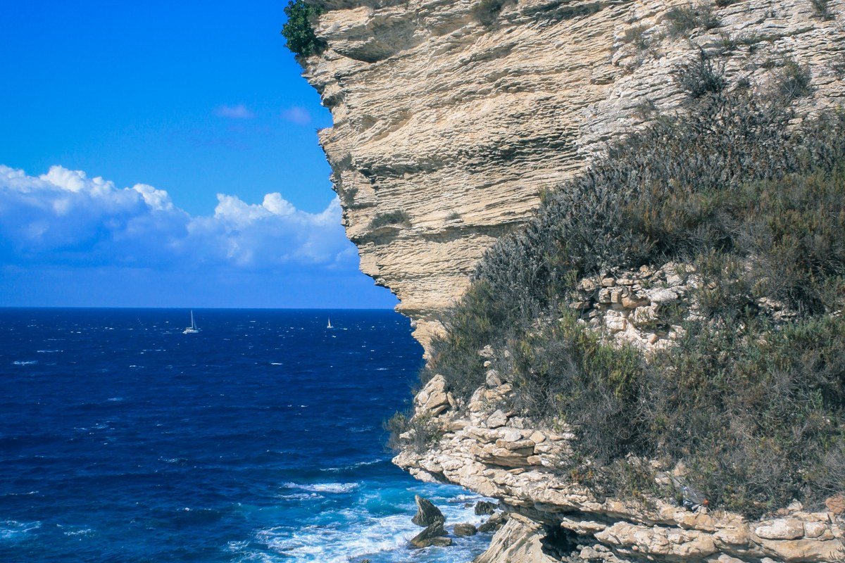 Bonifacio photographed from a boat on the strait, the old town appearing to overhang white limestone cliffs above cobalt water, Sardinia faintly visible on the horizon