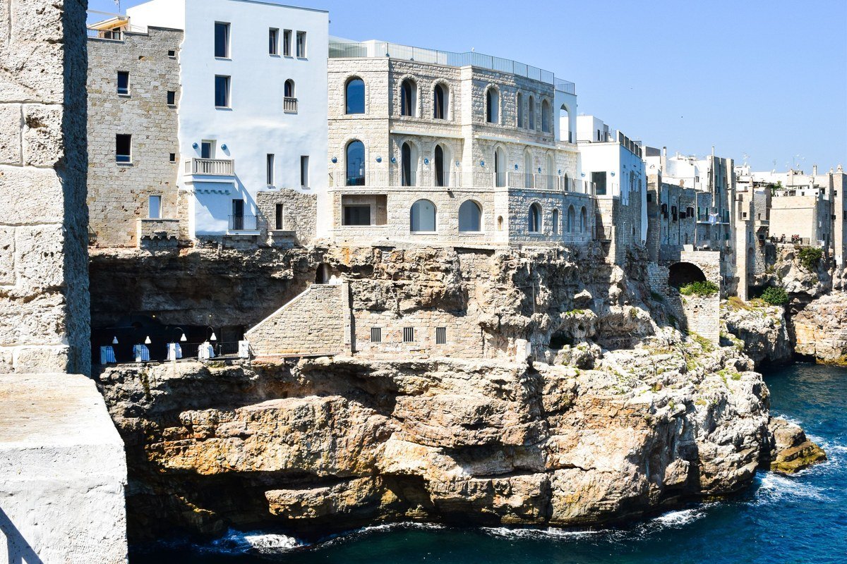 Polignano a Mare's cliff path at sunset, low white limestone above clear Adriatic water, the sea caves visible below and no restaurant tables required to justify the view