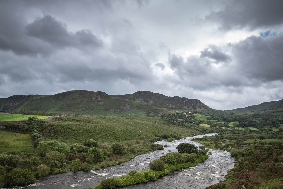 The River Coe near Altnafeadh after overnight rain, the water running dark peat-brown, low mist cutting the Three Sisters off at mid-height so only their lower flanks are visible. The immediate foreground, lichen-covered rocks and wet grass, is pin-sharp. Shot at 70mm to compress the mist layers. No filter. No composite. This is what the valley floor looks like thirty minutes after a heavy shower moves through.