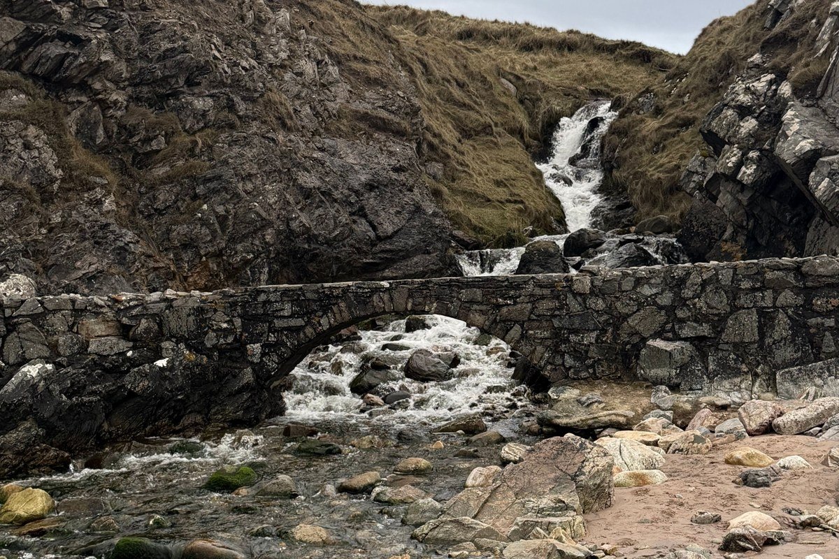 Sligachan Old Bridge from the downstream bank, the Cuillin Ridge half-hidden in rain cloud, the river running fast and heavily peated, the old stone arch framing a wall of mist where the mountains should be. Shot in October at 8 a.m., two minutes after a rain shower passed. The mountains reveal themselves in sections for about twenty minutes before the next band of cloud closes in. That twenty-minute window is the whole game here.