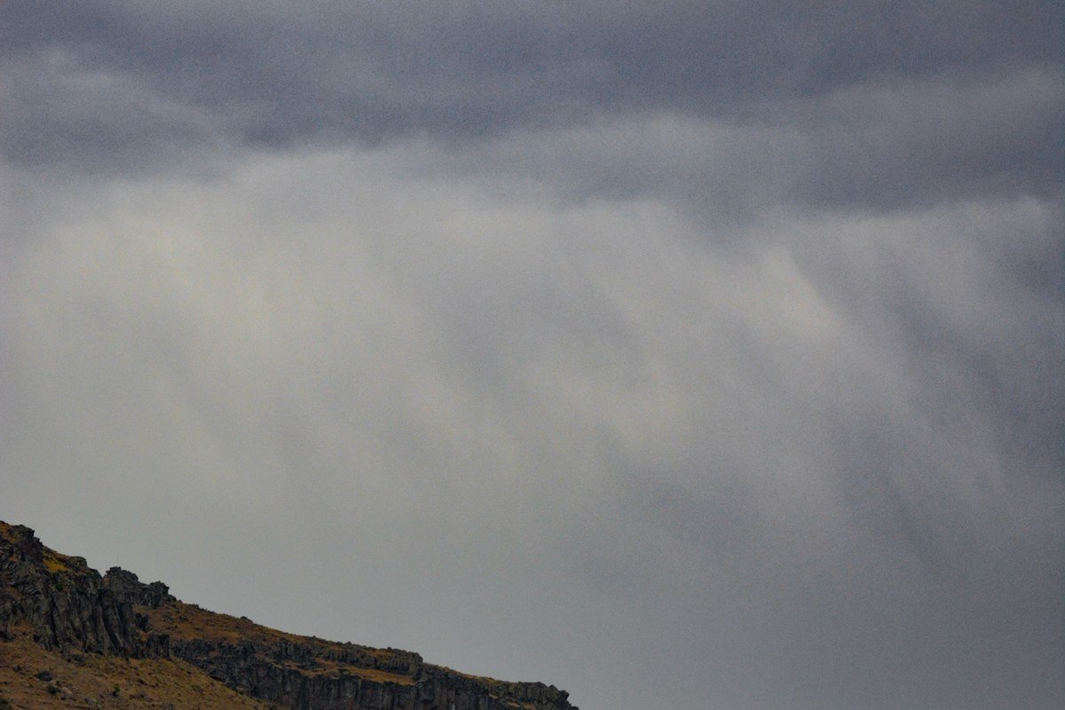 The upper hairpin bends of Bealach na Ba in low cloud, the road surface wet and reflective, a temporary waterfall running across the tarmac from the hillside above, the next bend invisible in mist twenty metres ahead. Shot from the car with the window open, engine off. No other vehicles in either direction. The pass was like this for forty minutes before the cloud lifted. Drive it slowly with the window down and you hear the water before you see it.
