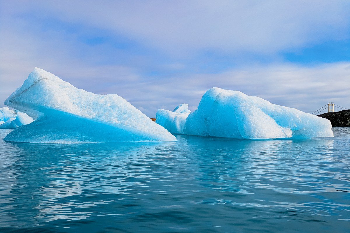 A sea kayak paddles beneath a blue ice arch inside Bear Glacier Lagoon, with the calved berg towering twice the kayak's length.